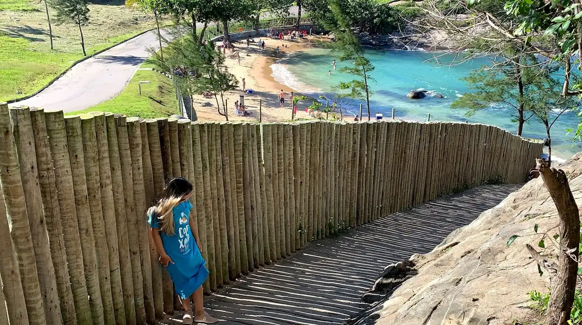 Mulher observando o mar turquesa em uma praia tranquila do Espírito Santo, com acesso por uma escadaria de madeira rústica.