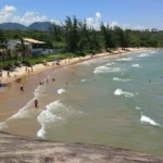 Vista da Praia dos Adventistas em Guarapari com castanheiras fazendo sombra natural na areia e mar calmo.