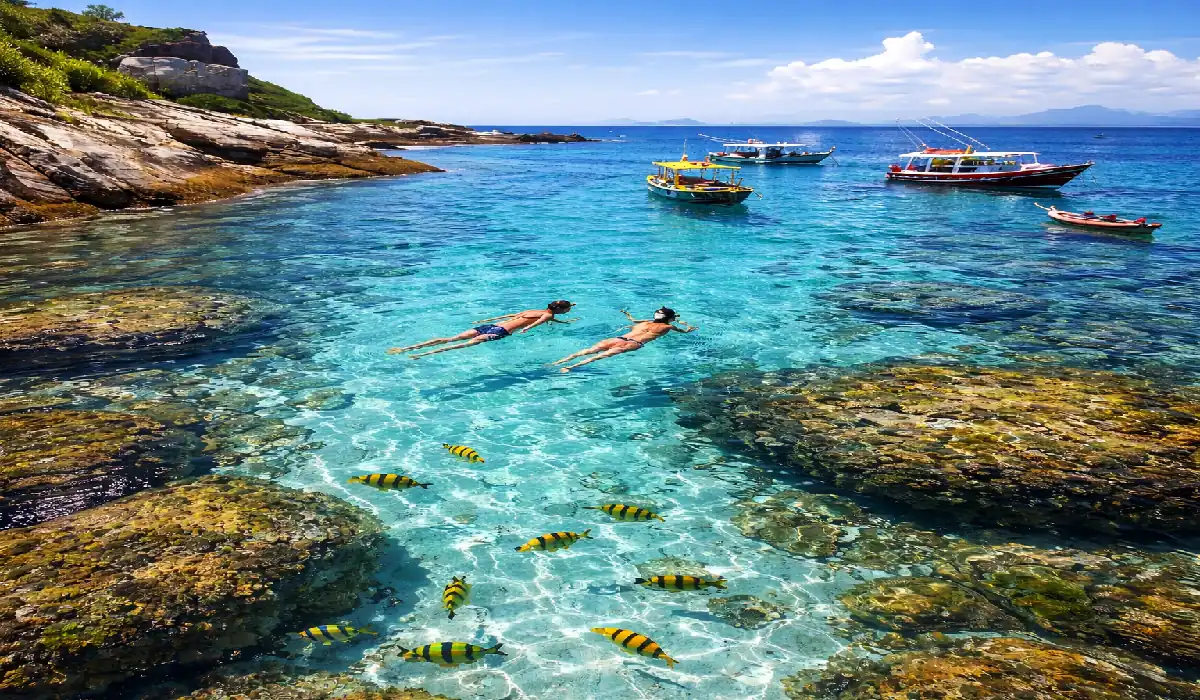 Mergulho em águas transparentes cor turquesa com peixes coloridos em primeiro plano e barcos ao fundo, representando as ilhas do Espírito Santo.