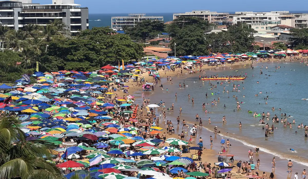 Vista panorâmica da Praia de Peracanga, em Guarapari, completamente lotada. A faixa de areia está coberta por centenas de guarda-sóis coloridos e muitos banhistas aproveitam o mar calmo da Enseada Azul em um dia de sol.